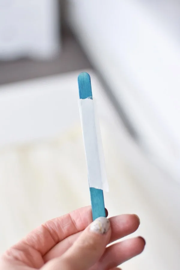 Woman holds blue popsicle stick with strip of fabric tape on it.