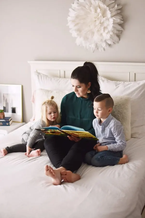 Brunette woman sits on bed reading a book while boy and girl listen beside her. 