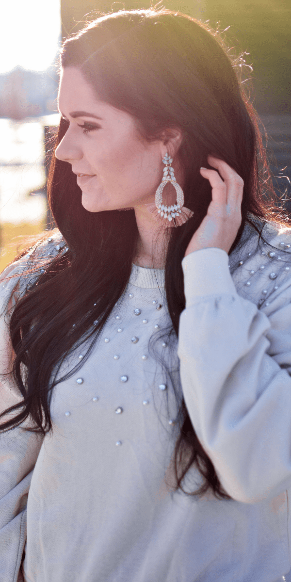 A Woman in a gray sweater with beading tucks her hair behind her ear to reveal statement earrings.