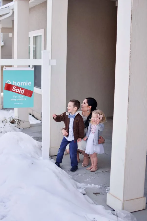 A mom and two kids stand next to their Homie sign in front of their sold house.