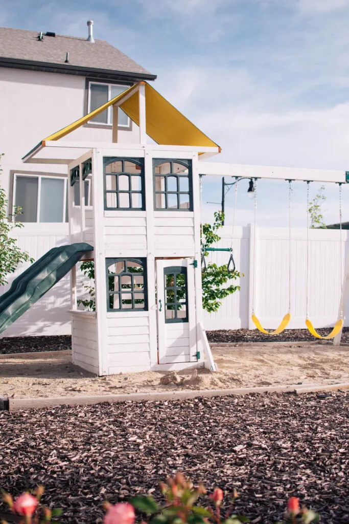 A white swing set with a tower surrounded by a sand box in a backyard.