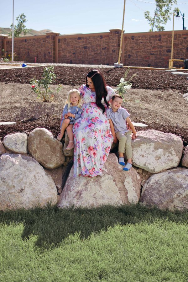 A mom sits with her two children on a large rock wall in their backyard.
