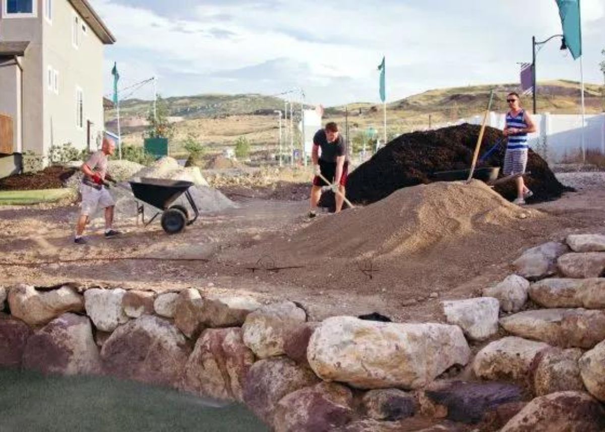 People working on a backyard landscaping project with piles of dirt, mulch, and wheelbarrows.