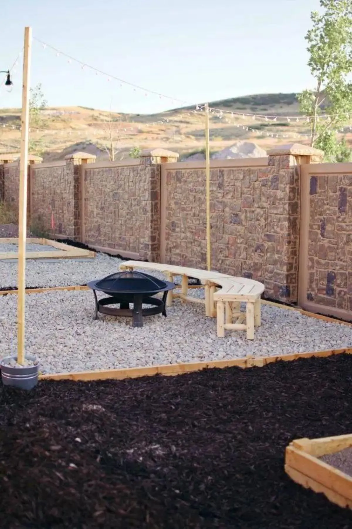 A fire pit with gravel and a wooden bench under string lights in a backyard with a stone fence.