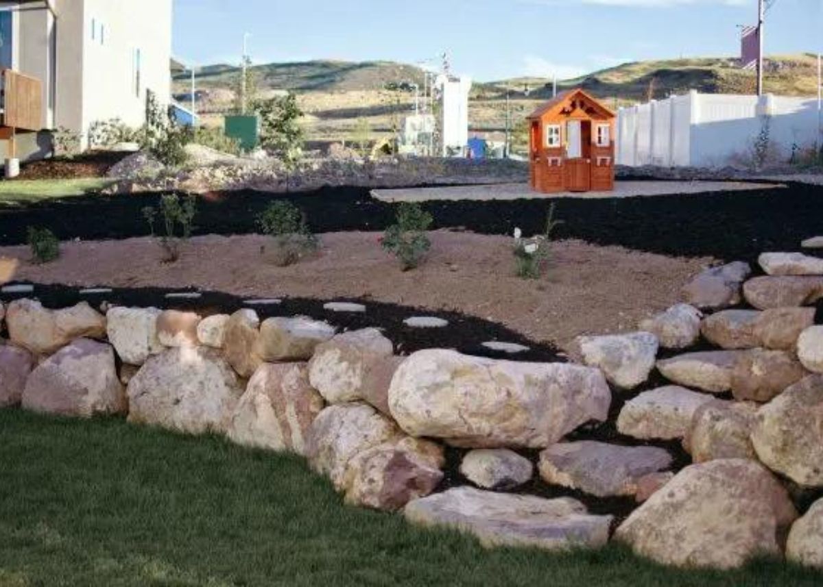 Boulders create tiered landscaping in a backyard with plants and a wood playhouse.