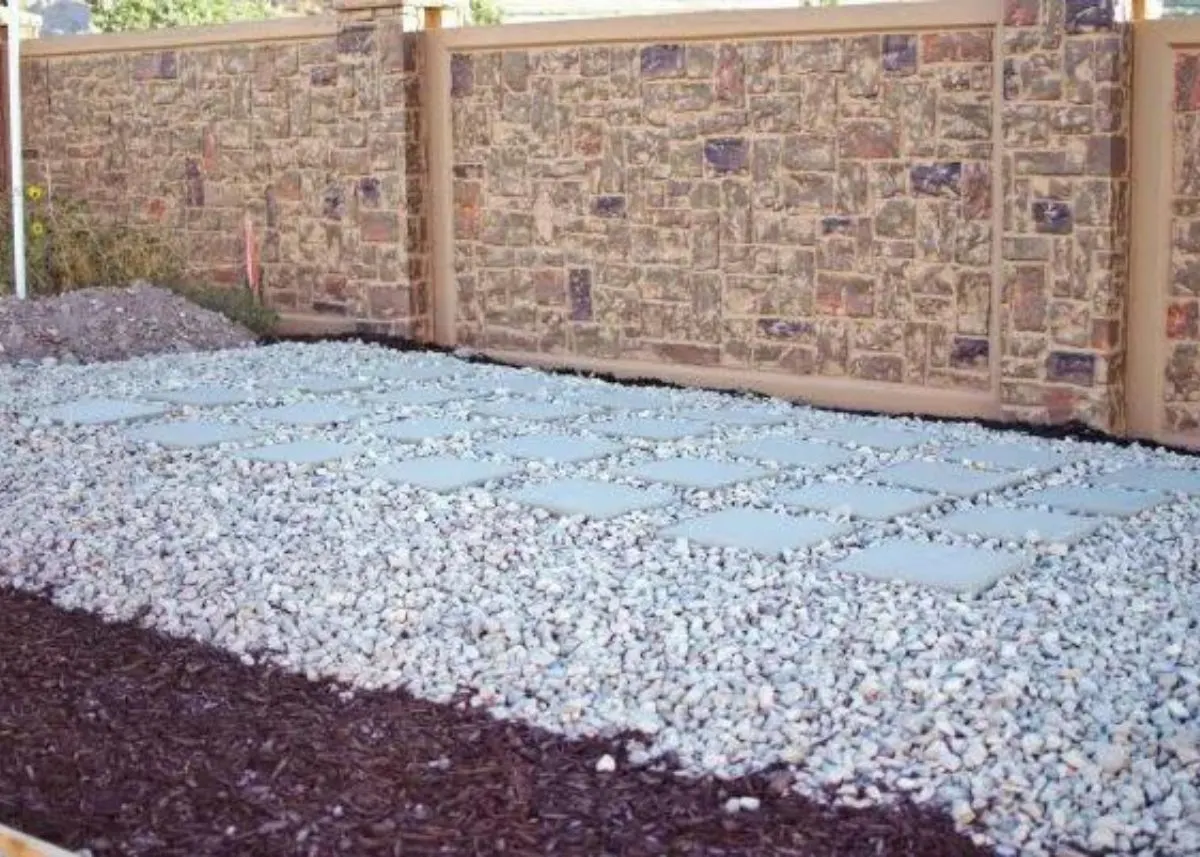 Gravel and dark mulch landscaping with cement stepping stones in front of a stone fence.