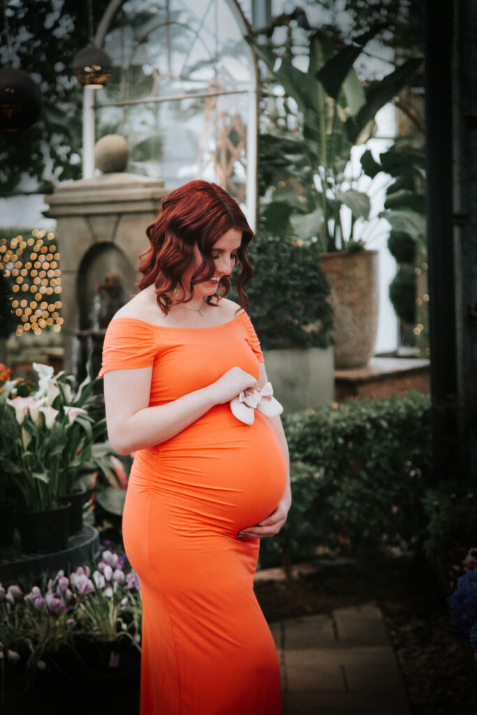 Woman wearing orange dress holds baby shoes on stomach during greenhouse maternity pictures.