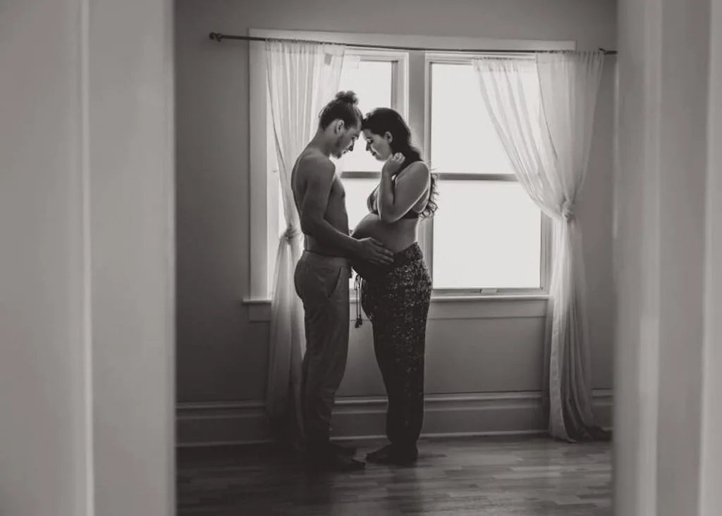 Husband touches wife's baby bump next to a window during maternity photo shoot.