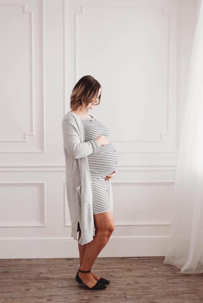 Woman wearing striped dress and cardigan holds bump during studio maternity pictures.