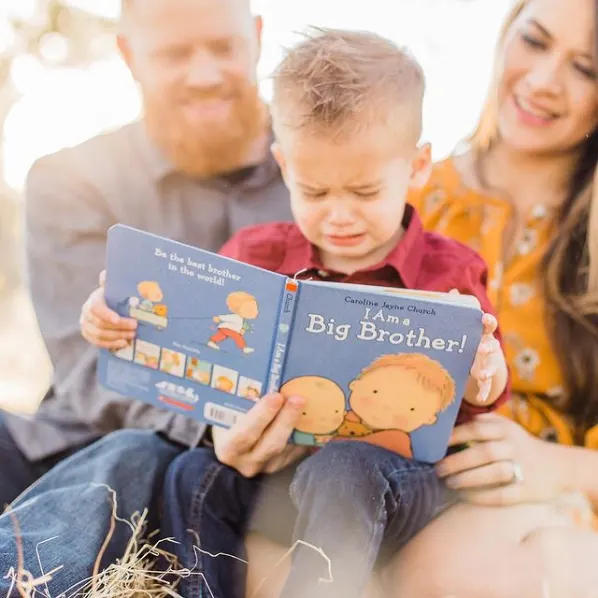 Little boy sits on his mom and dad's lap and holds a book about becoming a big brother.