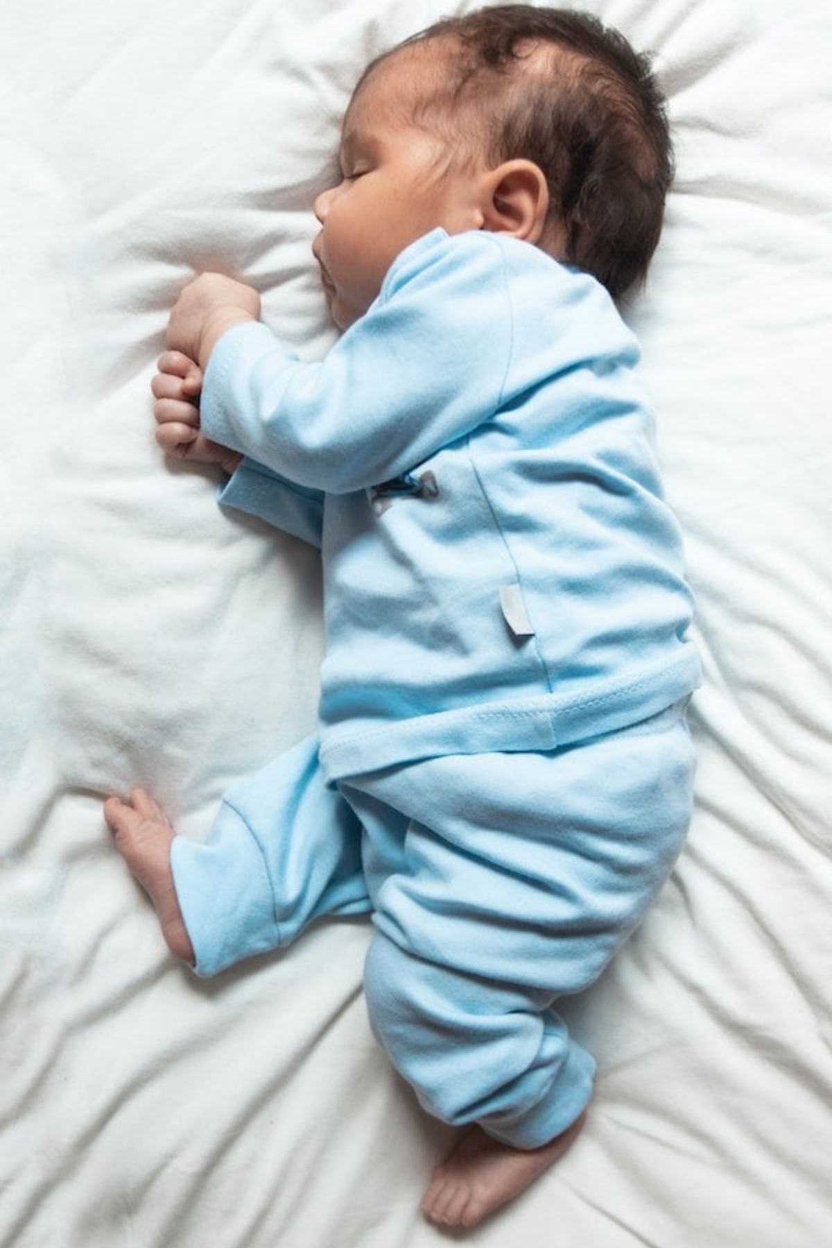 Baby boy wearing blue outfit lays on side while sleeping on white sheet.