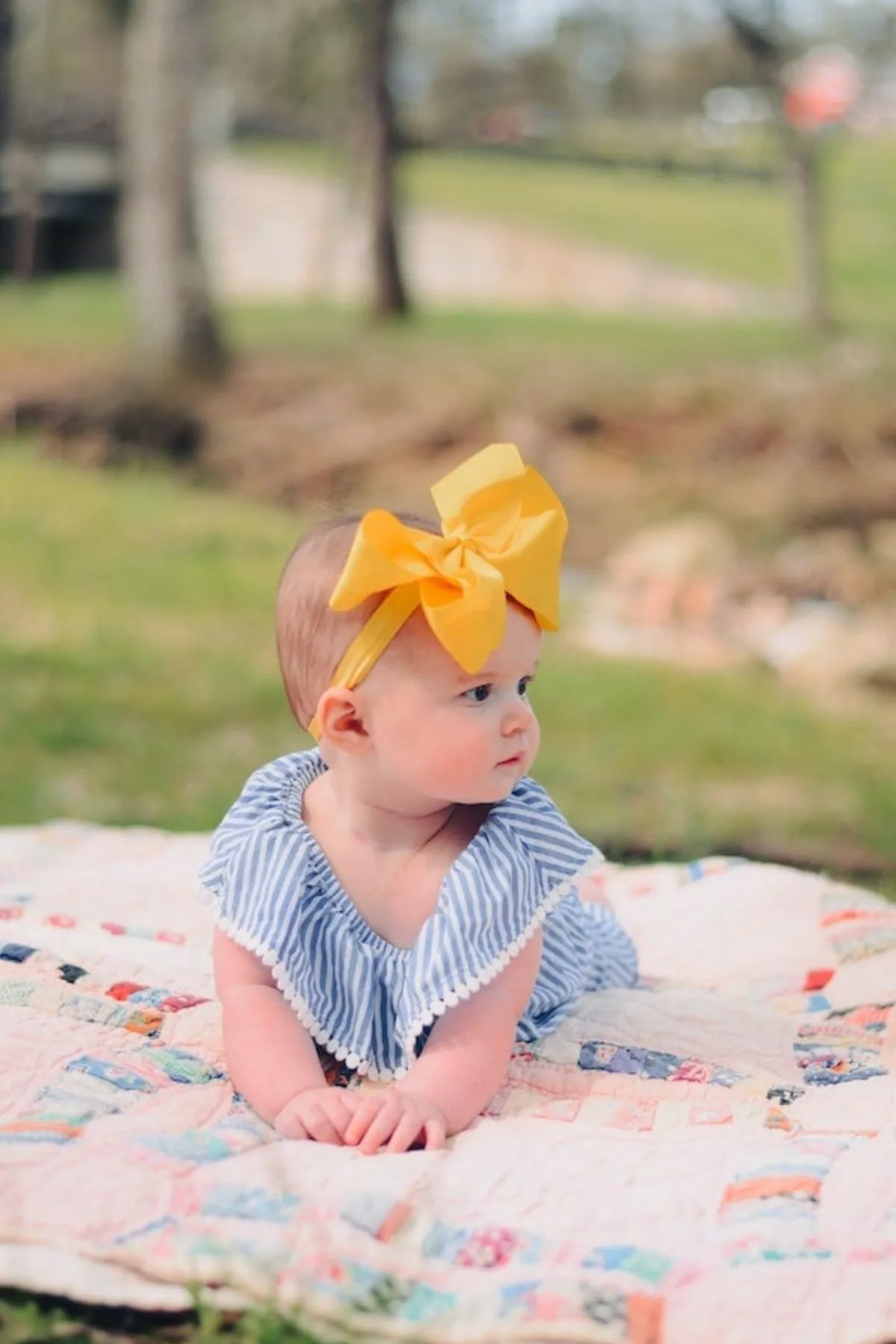 Baby girl with yellow bow and blue striped romper pushes up on patchwork quilt outside.