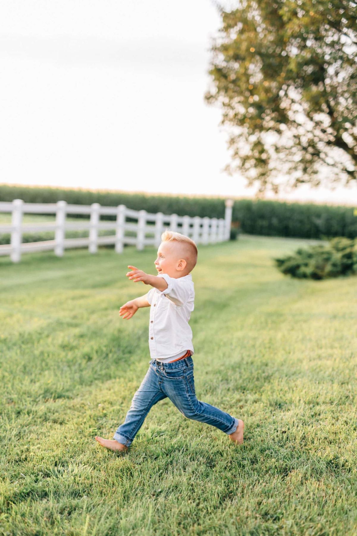 Toddler boy wearing white button up and jeans runs on a lawn while laughing.