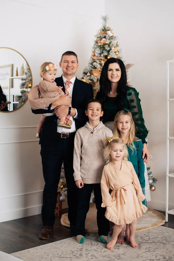 Family of 6 wearing green and beige dress clothes stand in front of Christmas tree.
