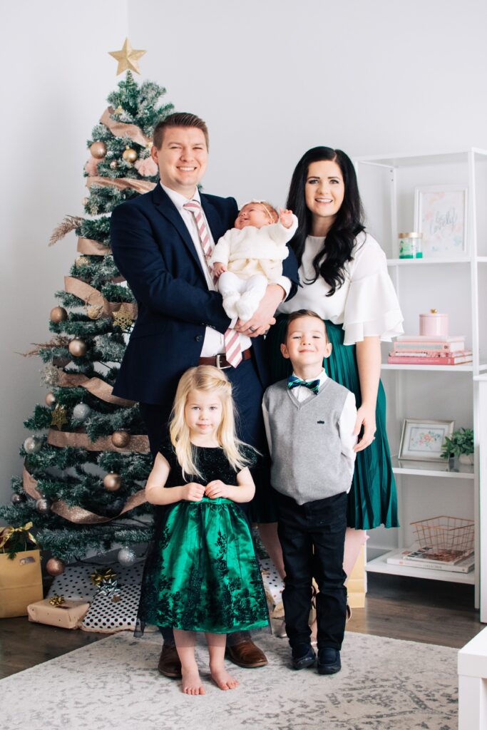Family of 5 wearing green dress clothes stand in front of Christmas tree.