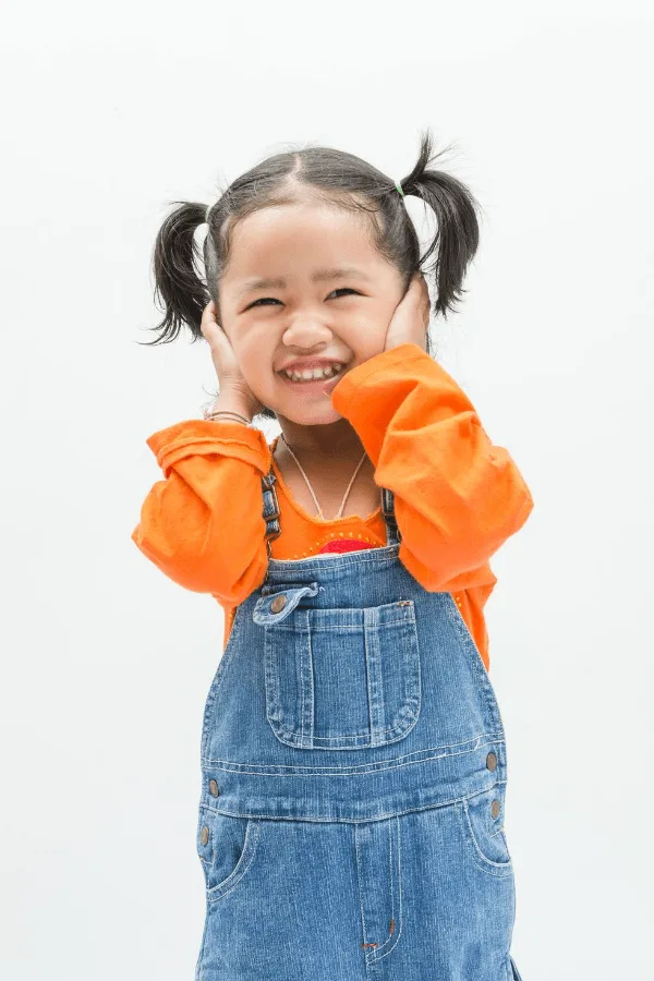Little girl with brown hair in pigtails puts her hands on her cheeks and smiles. 