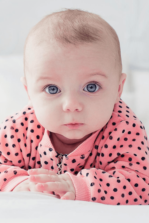 Baby girl with blue eyes wearing pink and black polka dot jacket looks at the camera.