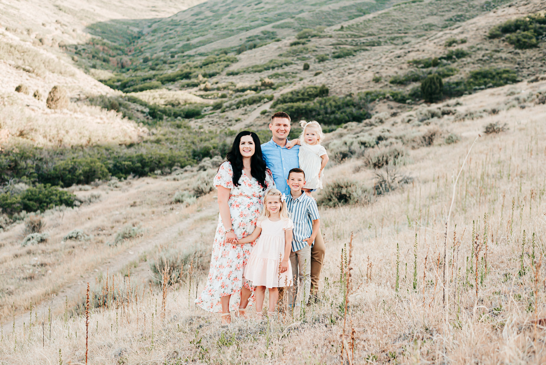 Family of 5 wearing peach and blue outfits smiles on grassy mountain side for a family photo.