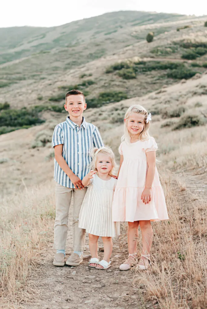 Three kids wearing blue, cream, and peach outfits smile on grassy mountainside for a family picture.