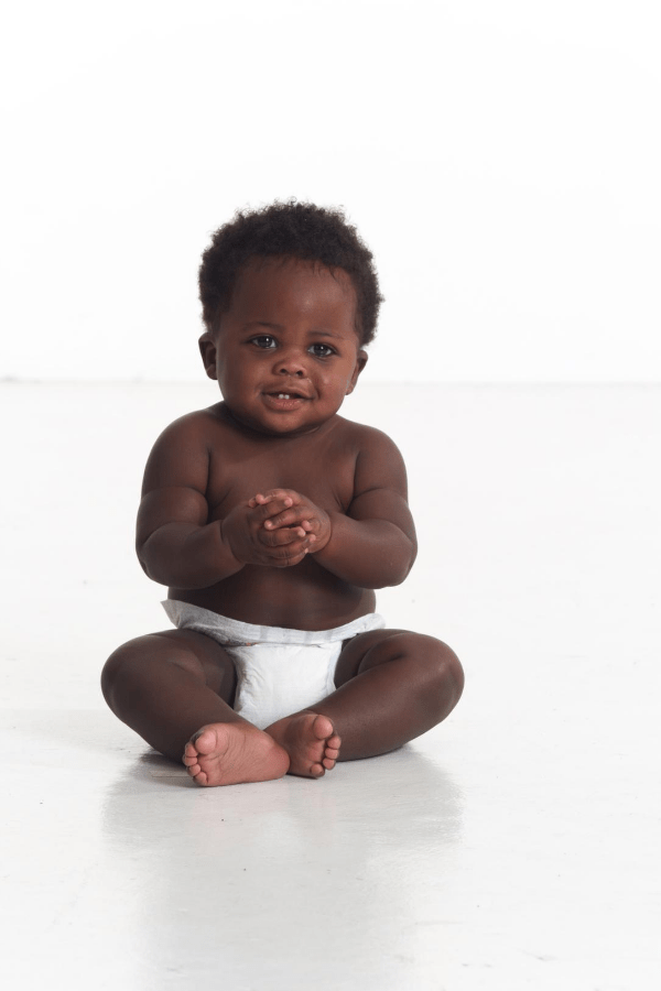 Black baby boy wearing white diaper sits on a floor while holding hands together.
