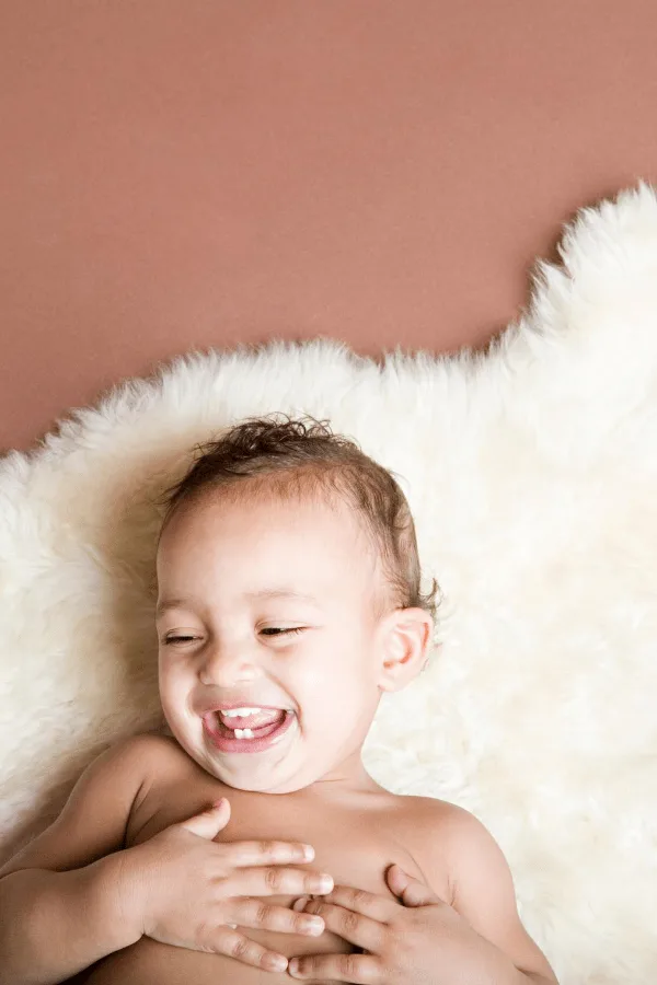 Baby boy with brown hair lays on a white rug and laughs.