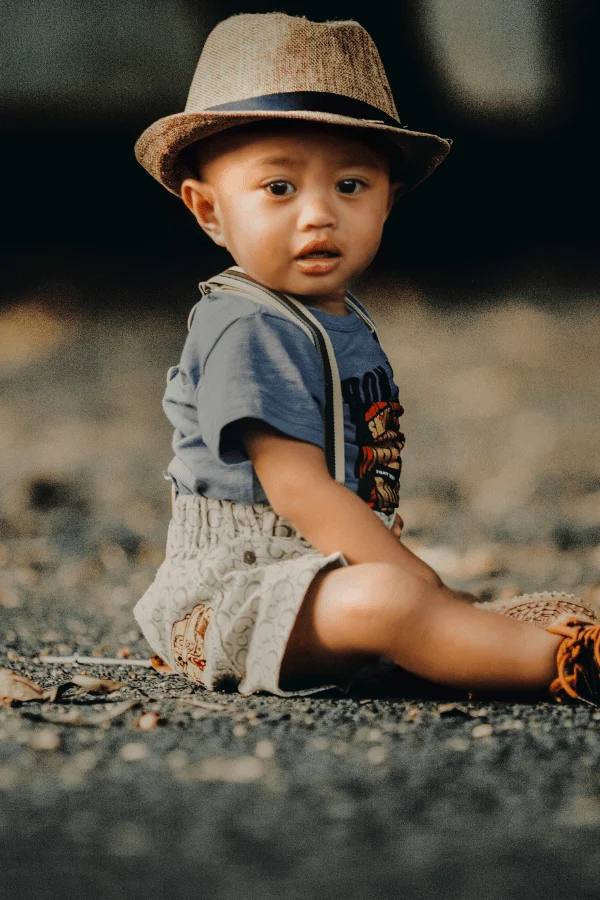Baby boy sits on the ground wearing a hat and suspenders.