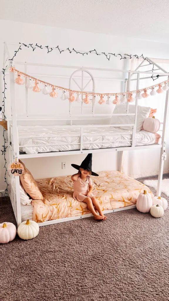 Little girl wearing black witch hat sits on bottom bunk bed decorated for Halloween.