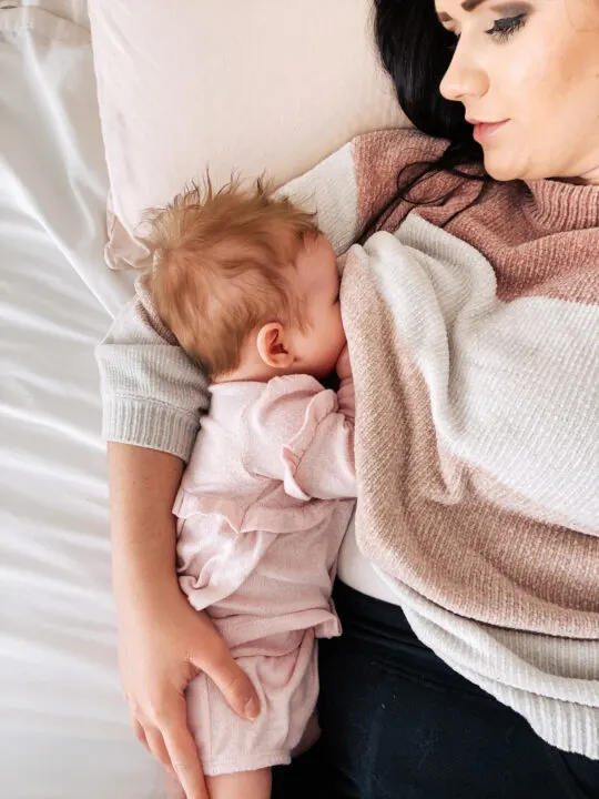 Mom breastfeeds her baby girl while lying on a bed with pillow.