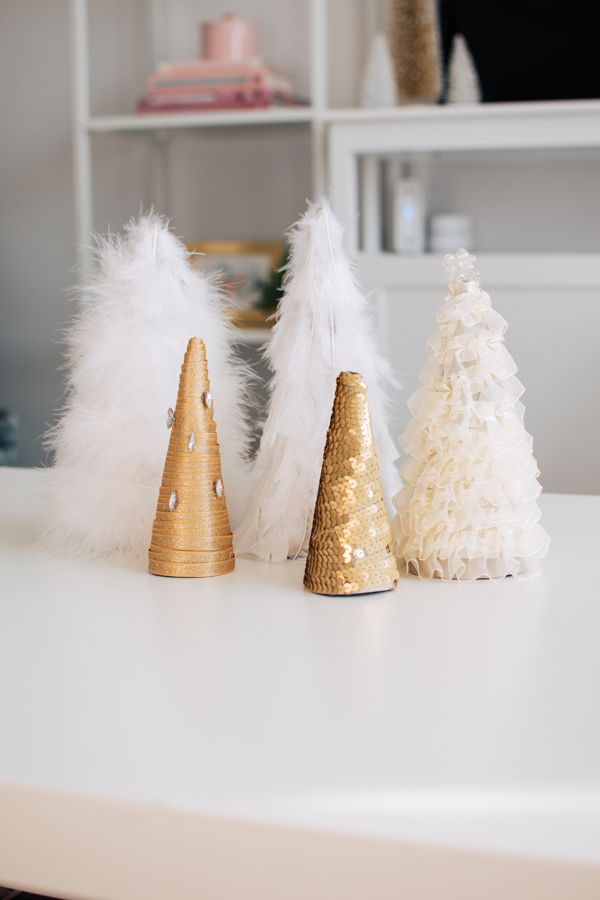 DIY cone Christmas trees decorated with feathers, sequins, ribbon, and jewels on white table.