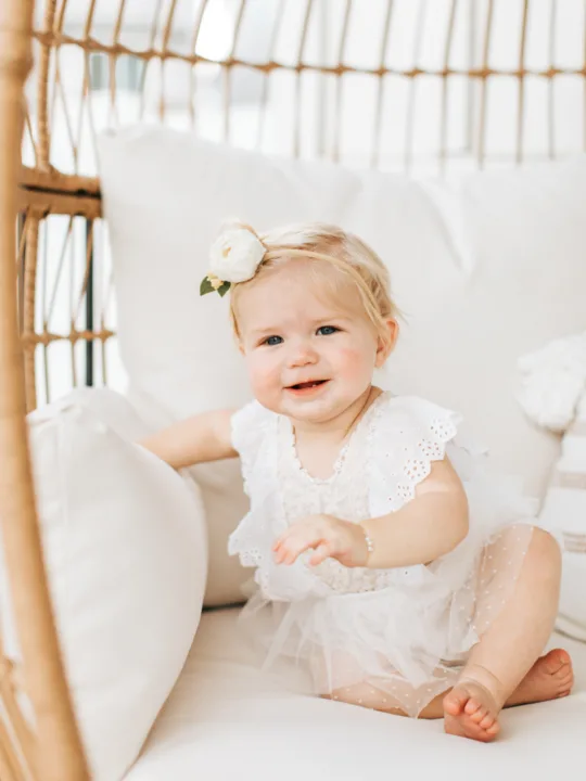 Baby girl wearing a white outfit sits on a chair.