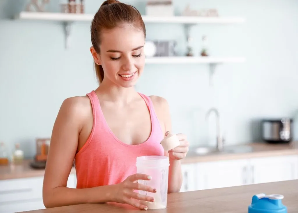 Woman wearing pink tank top stands in kitchen and pours protein powder into a blender bottle.
