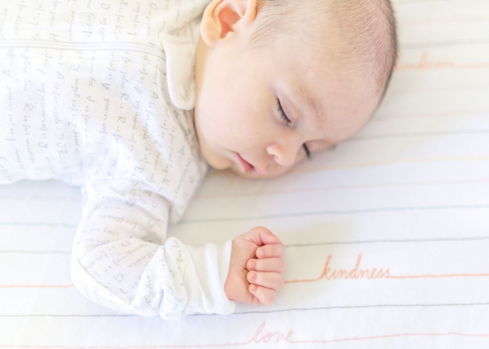 Baby wearing white outfit naps on sheets that say kindness.