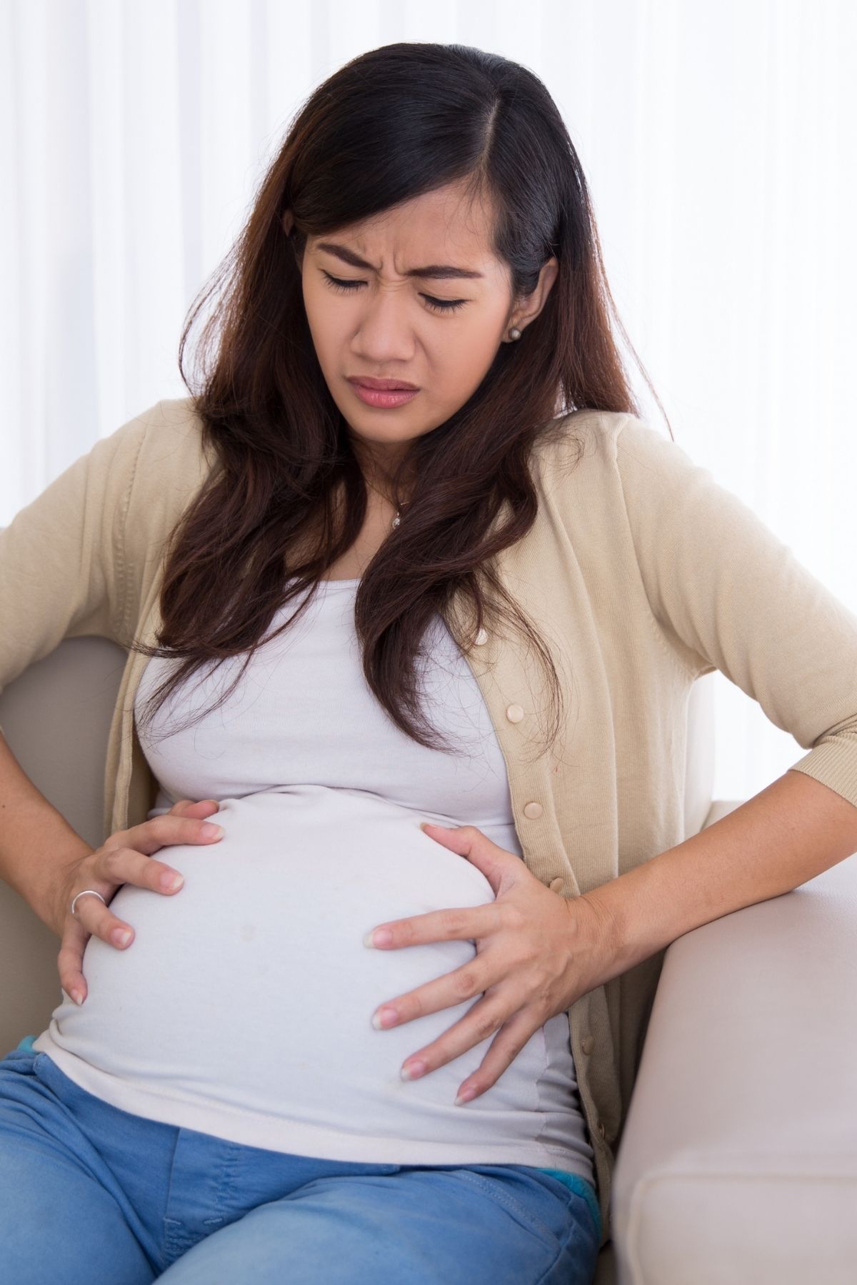Woman sitting on couch holds her pregnant belly while wincing from stomach pain.