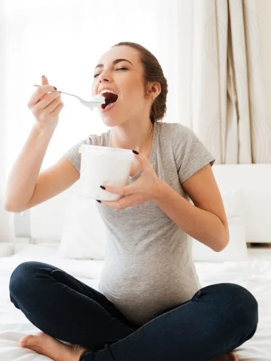 Pregnant woman eating ice cream from a tub on her bed.