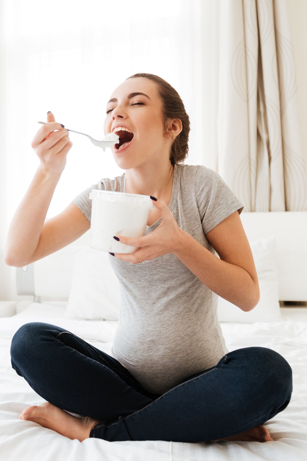 Pregnant woman sits on bed and holds a tub of white cream in one hand and a spoon with the cream on it in another.