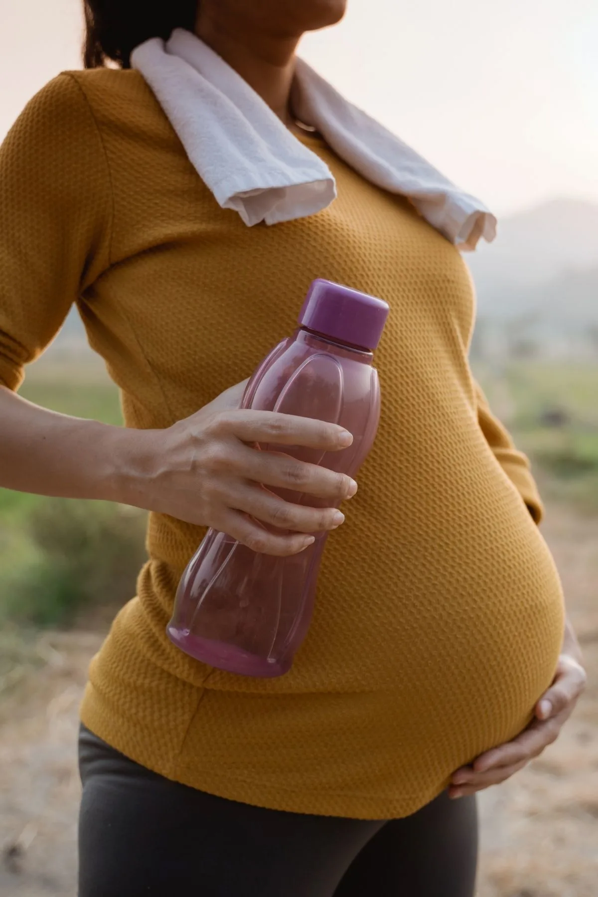 Pregnant woman holds water bottle and cradles baby bump while resting during a hike.
