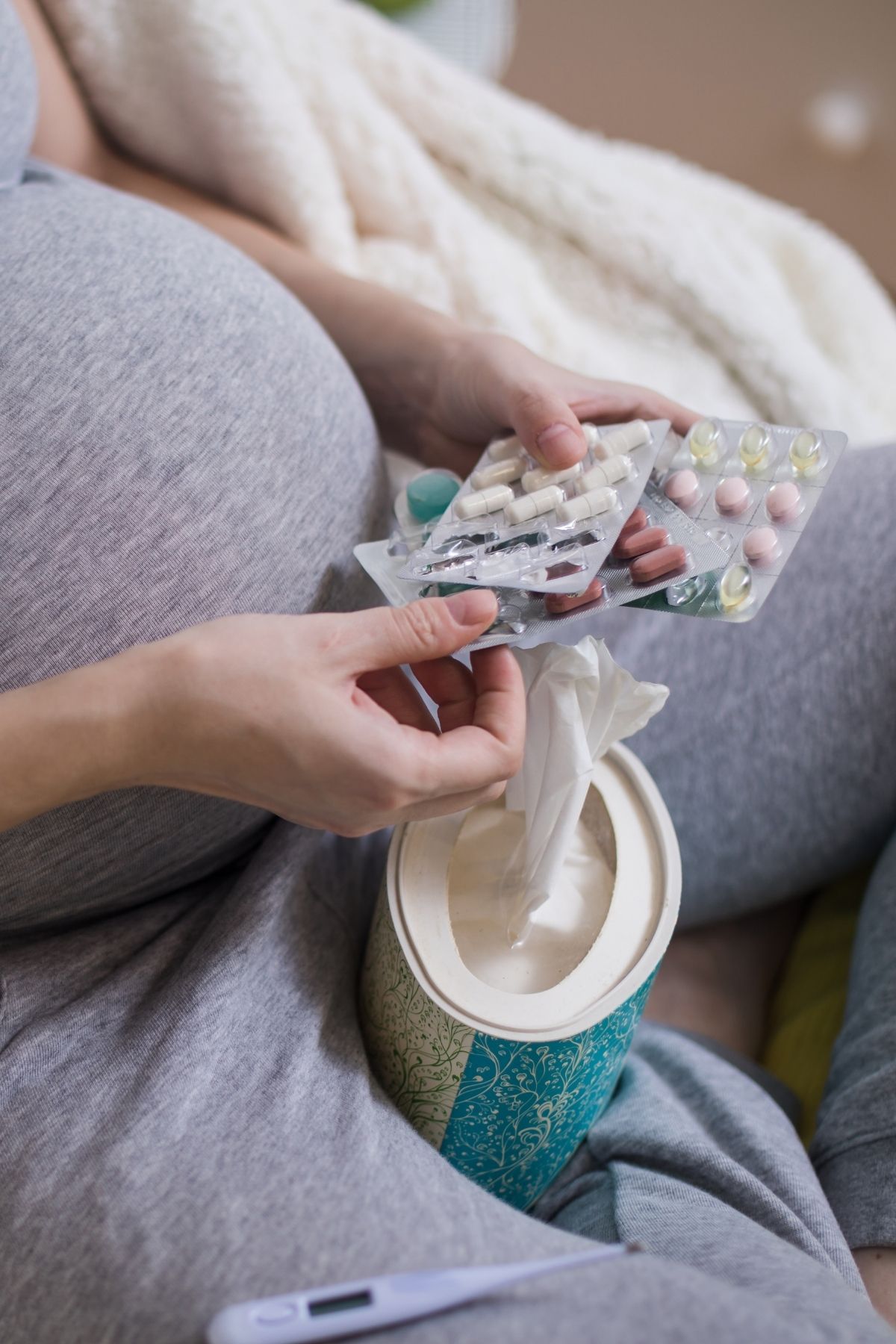 Pregnant woman holds several opened medication blister sheets while sitting cross-legged with a box of tissues.