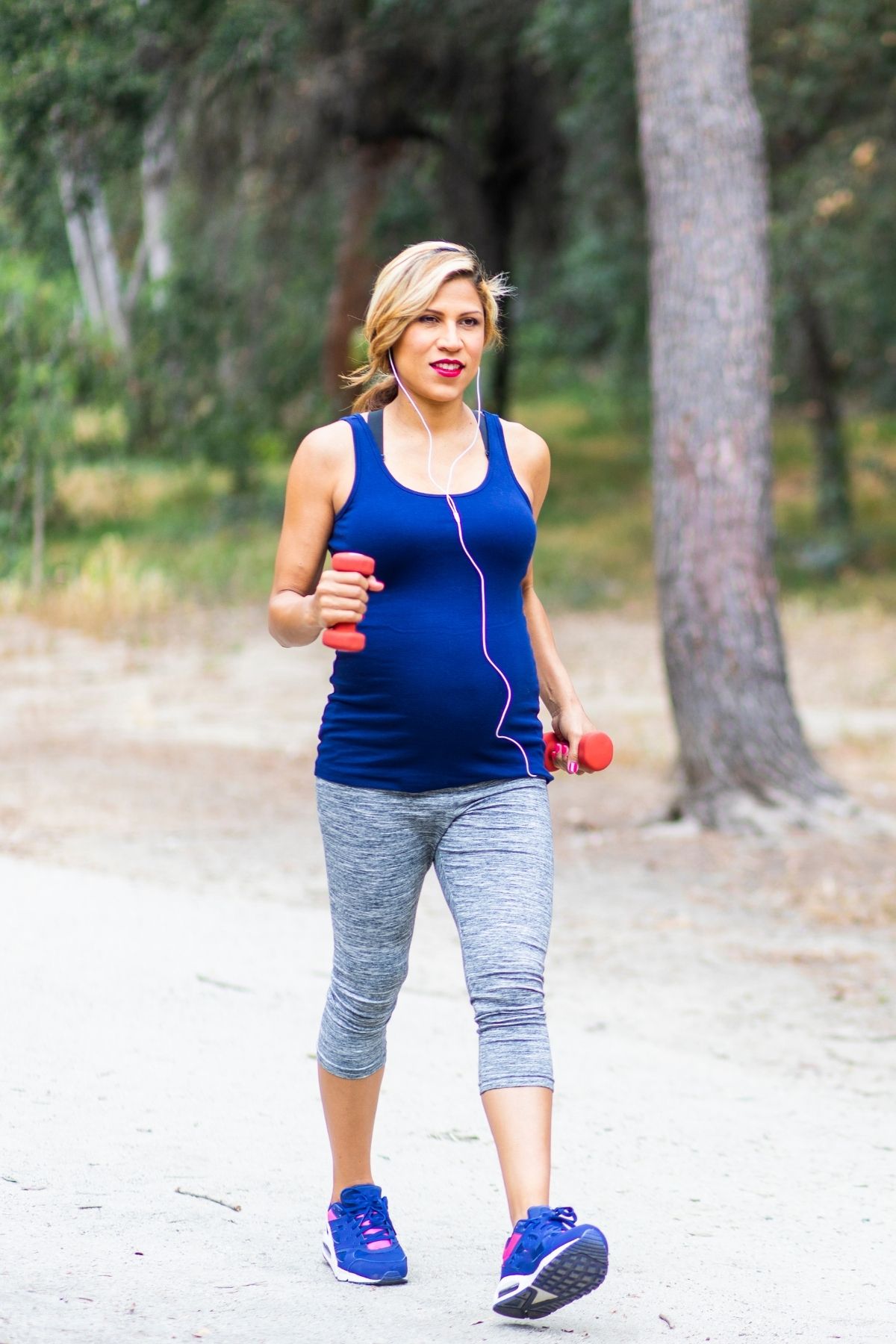 Pregnant woman uses weights and listens to music while walking outside.