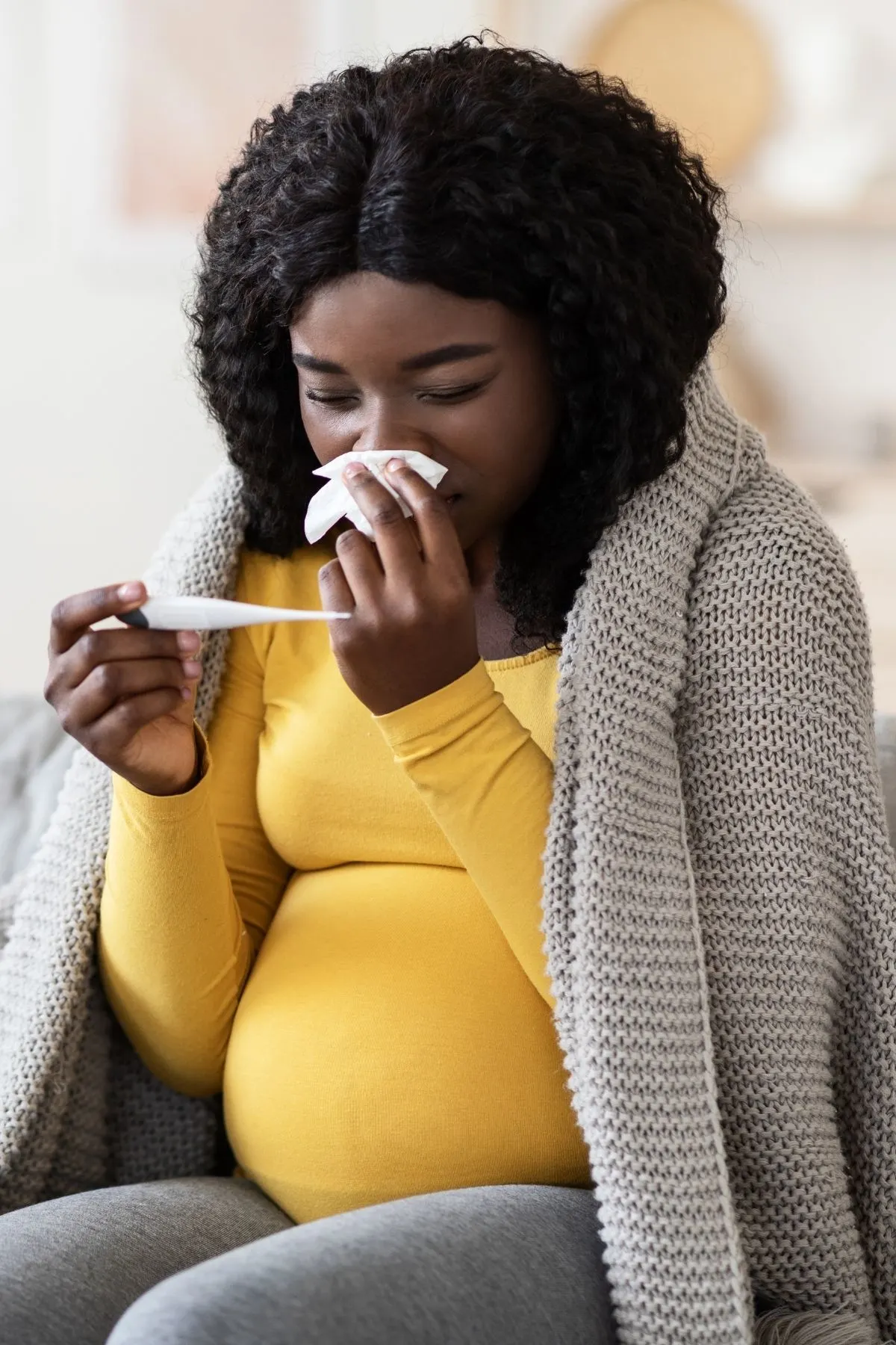 Pregnant woman with blanket over back blows her nose and checks temperature.