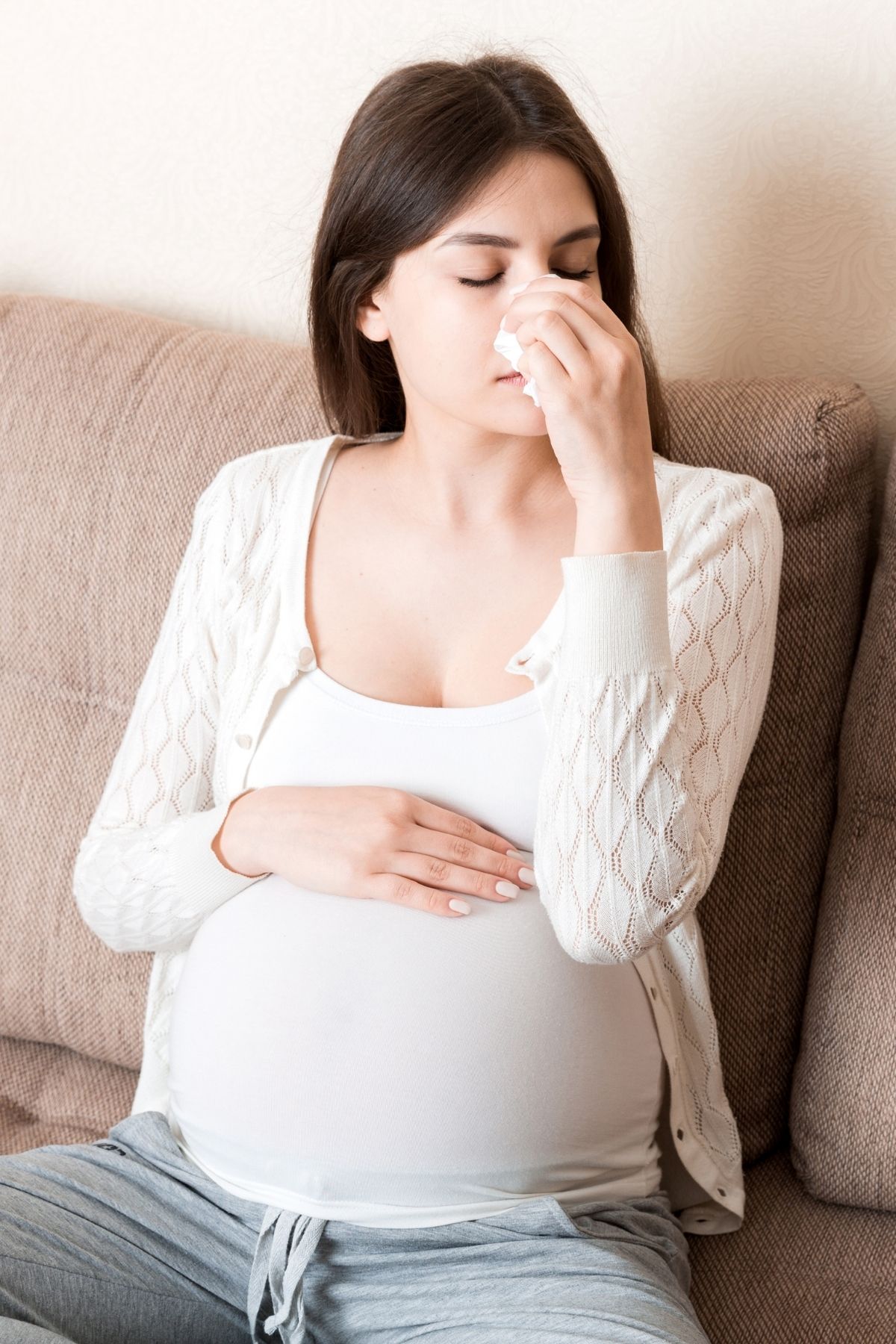 Sick pregnant woman rests hand on belly and blows her nose while sitting on couch.