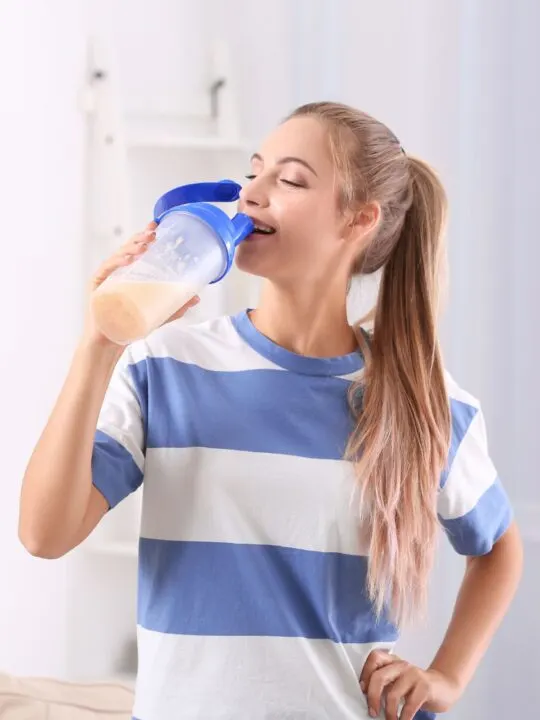 A woman in a blue and white striped shirt drinks a protein shake from a clear container.
