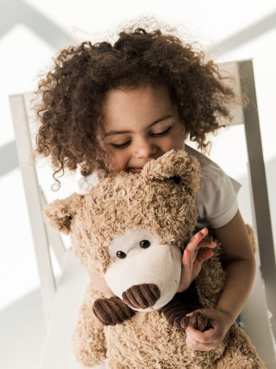 Girl with curly brown hair hugs teddy bear.
