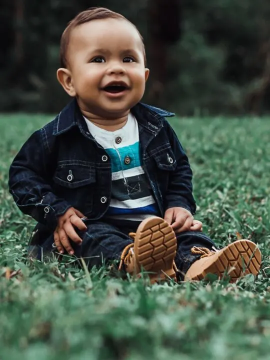 Baby boy in denim jacket and boots sits on grass.