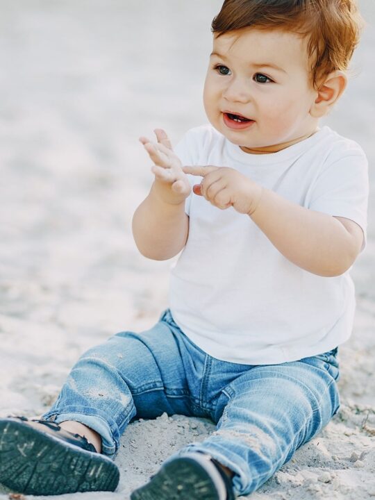 Baby boy sits on sand at the beach.