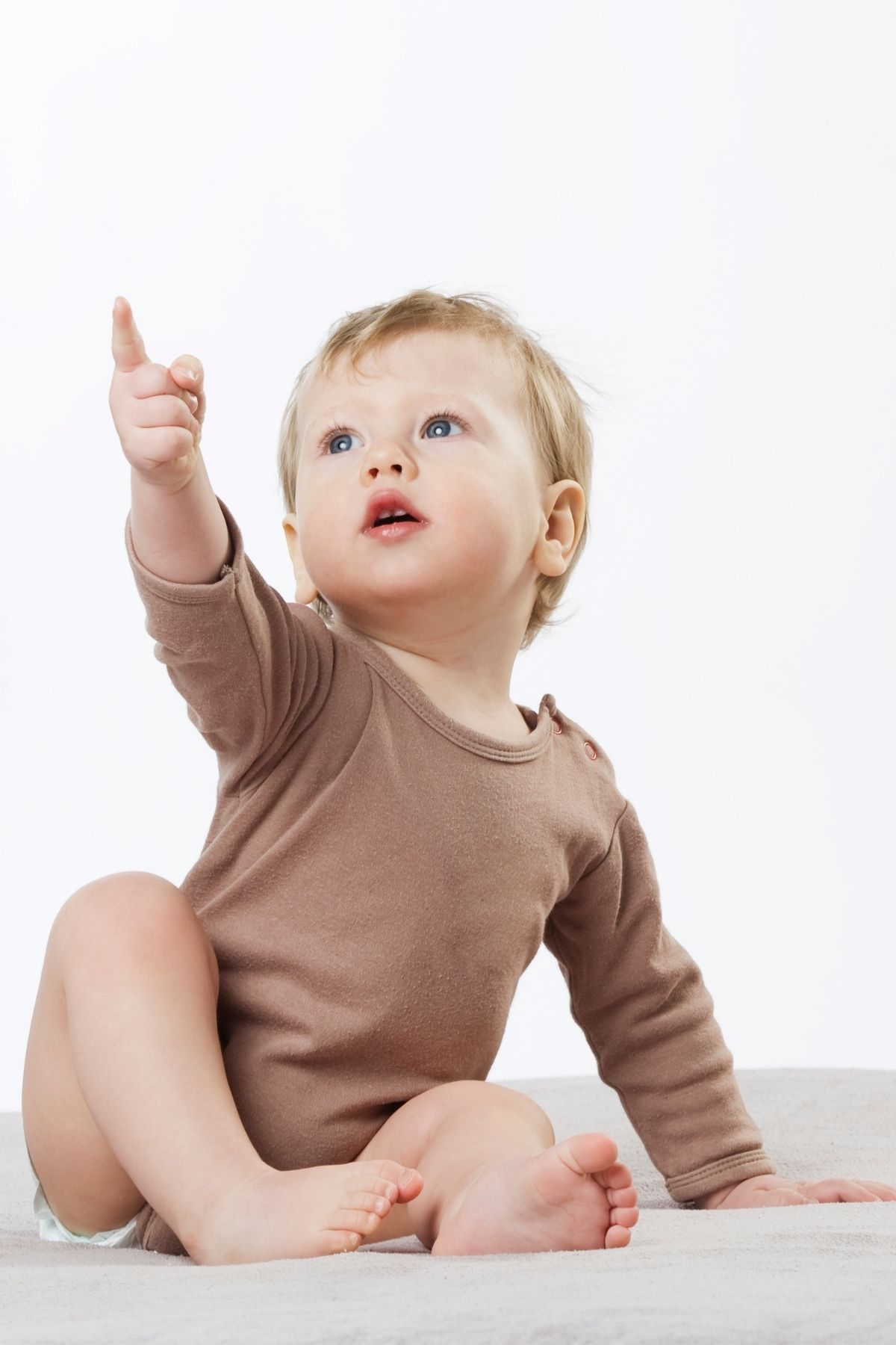 Baby boy wearing brown onesie sits in front of white background and points upward.