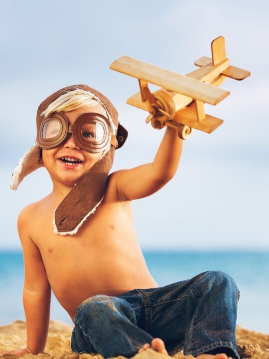 Boy plays with wooden airplane on beach.