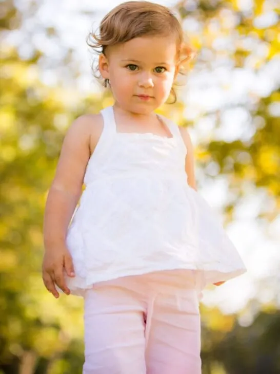 Toddler girl with brown curly hair wearing white shirt and pink pants stands outside.