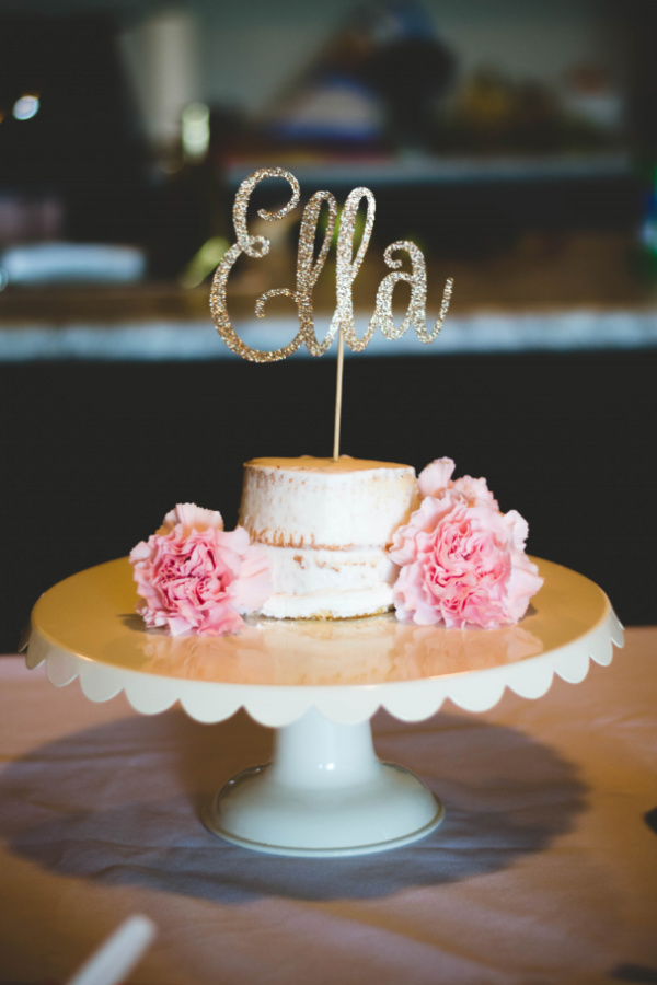Small birthday cake with gold lettered sign and pink carnations on white cake stand.
