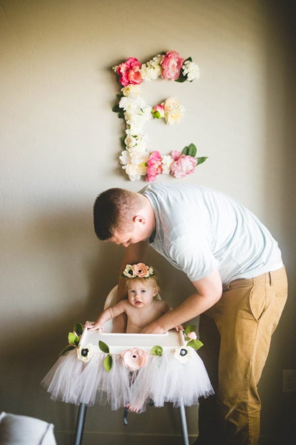 A little girl in a high chair decorated with flowers under a flower letter "E" with her father leaning over her.