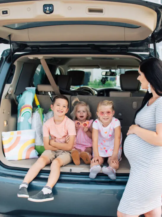 Three kids wearing sunglasses sit in trunk of car with summer items and smile while pregnant mom stands nearby.